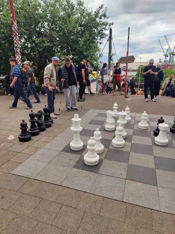 Eine Gruppe von Menschen unterschiedlichen Alters steht gemeinsam an einem überdimensionalen Schachbrett an einer Hafenpromenade